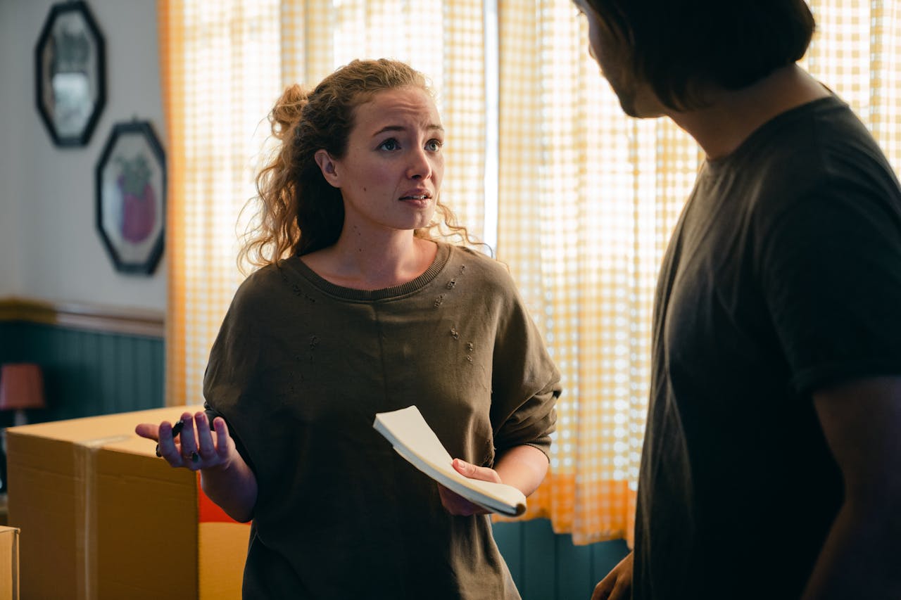 A young woman in a green sweatshirt holds a notebook in one hand and gestures with the other. Her hair is pulled back and she is talking to a man. They stand in a living room with moving boxes.